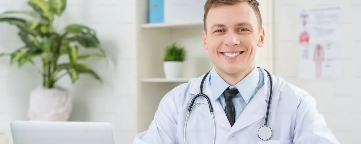 A general practitioner working at a consulting room desk with a laptop and patient notes