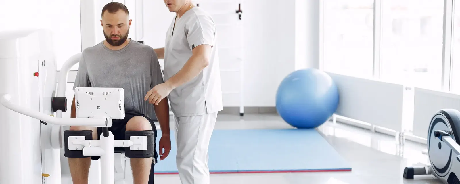 A patient using rehabilitation equipment with a practitioner in a bright exercise and rehabilitation space